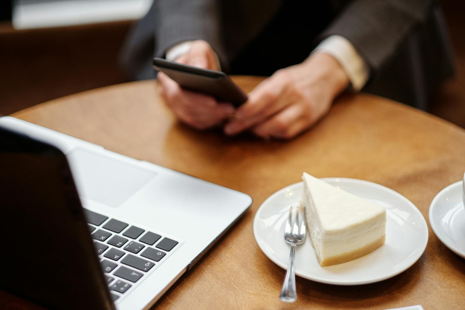 Businessperson using smartphone with laptop and cake in a cafe setting, perfect for work or lifestyle themes.