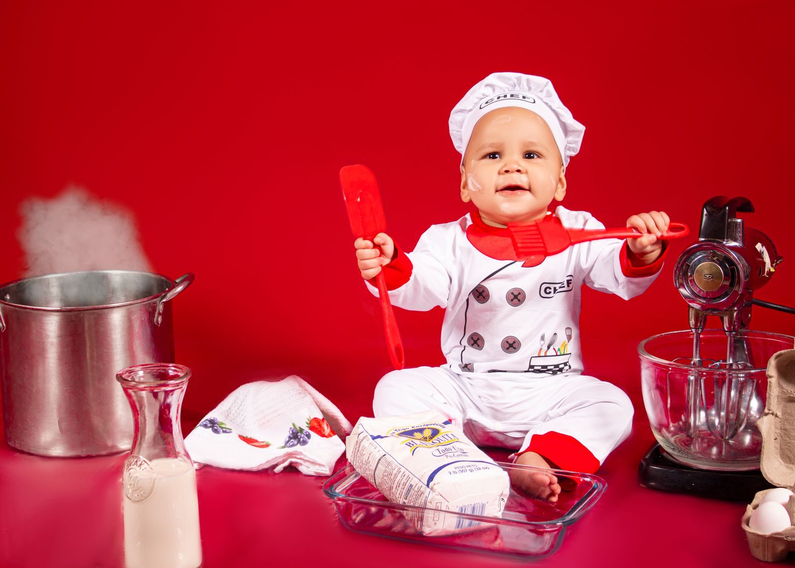 Cute baby dressed as chef playing with kitchen tools in vibrant red setting.